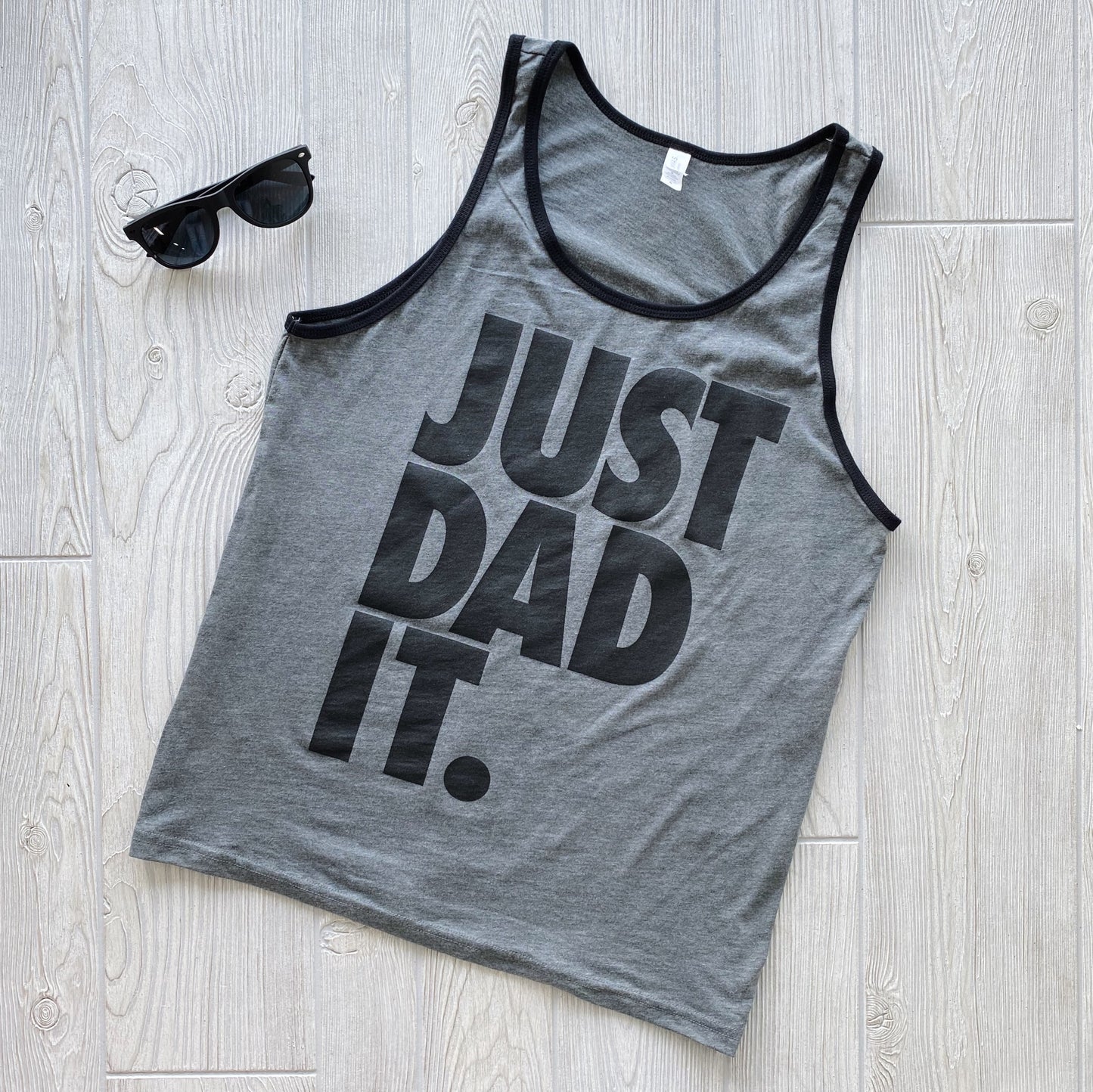 The Just Dad It • Ringer Tank is a gray ringer tank top with black trim and bold black JUST DAD IT text, shown beside black sunglasses on a light wood floor.
