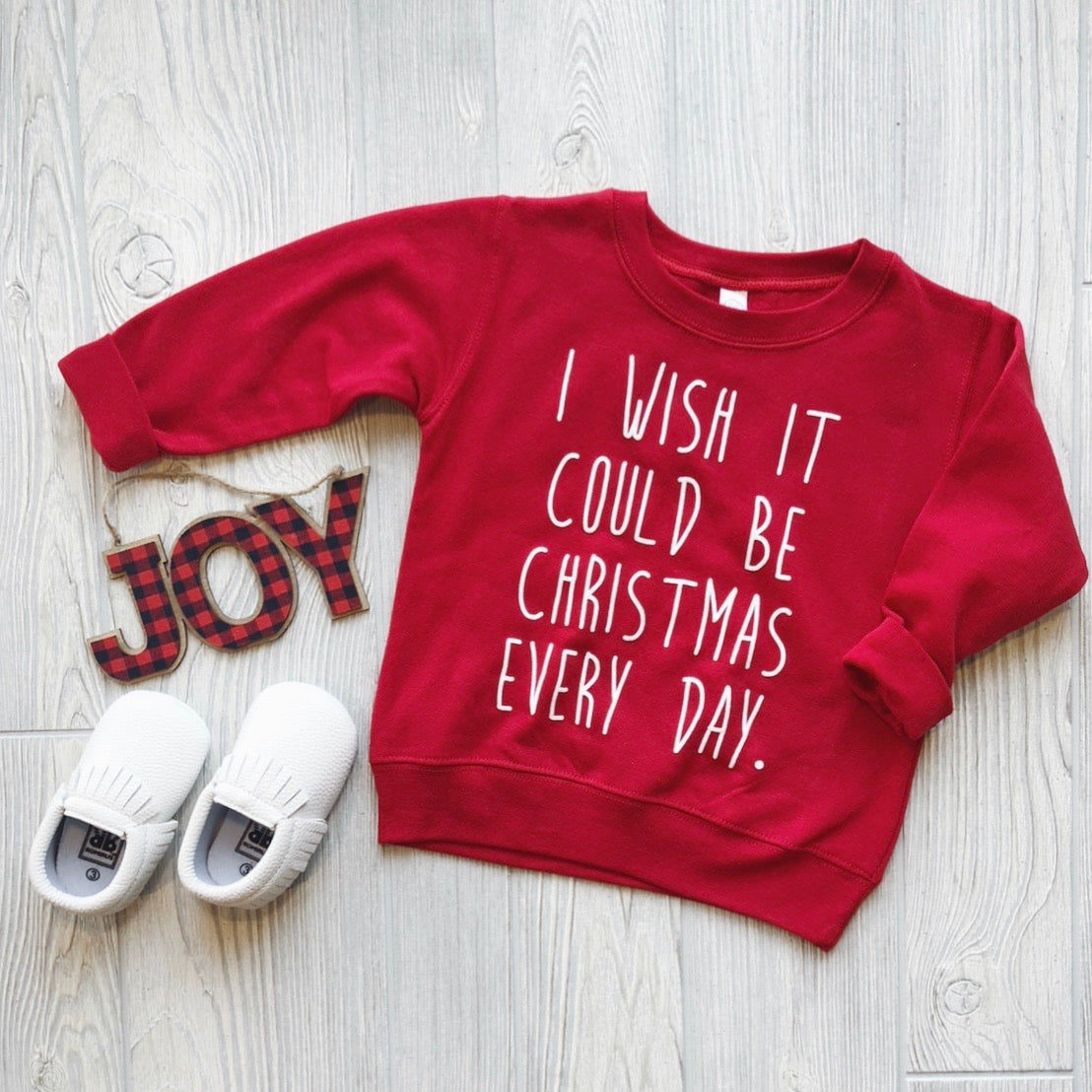 The Christmas Wish • Pullover, a red kids shirt with I wish it could be Christmas every day, is displayed on light wood flooring beside white slip-on shoes and a plaid JOY decoration—ideal festive holiday wear for little ones.