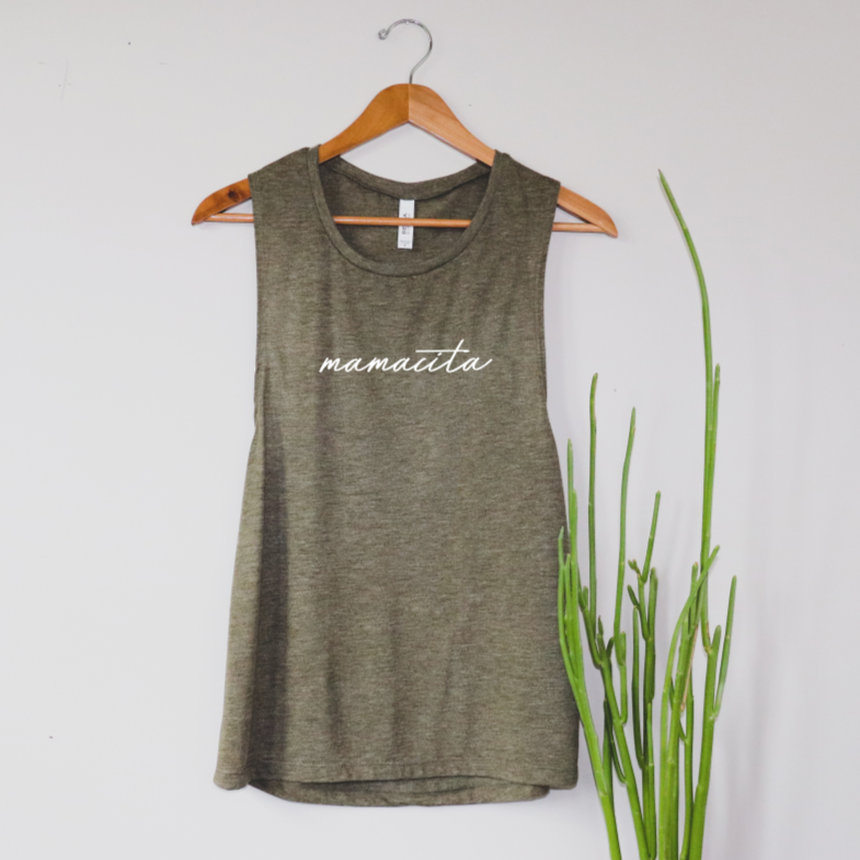 The Mamacita • Scoop Tank is a sleeveless olive green top with a relaxed drapey fit and white mamacita script, displayed on a wooden hanger next to a tall green plant against a plain white wall.