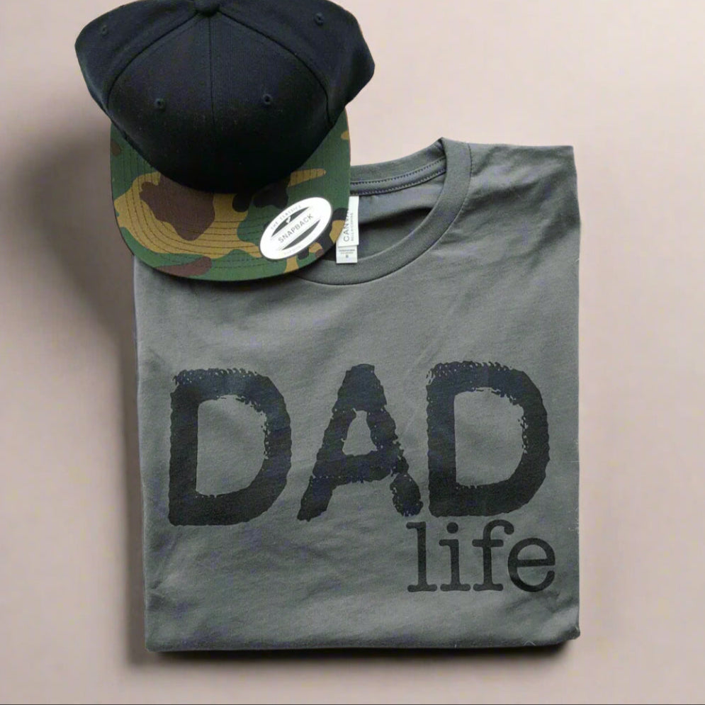 The Dad Life • Charcoal/Black set features a combed, ring-spun cotton T-shirt with “DAD life” in black and a black-camouflage baseball cap, displayed on a light surface.