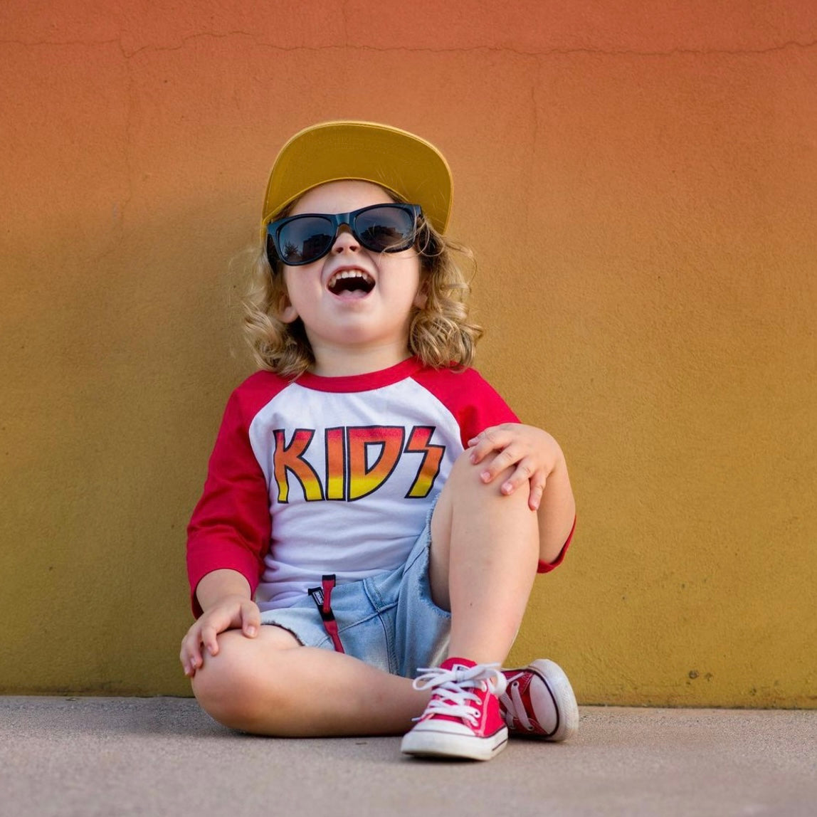 A young child with curly hair smiles while wearing a yellow cap, black sunglasses, denim shorts, red sneakers, and a true-to-size KIDS • Raglan shirt against an orange-yellow wall, showing off a cool little rocker style.