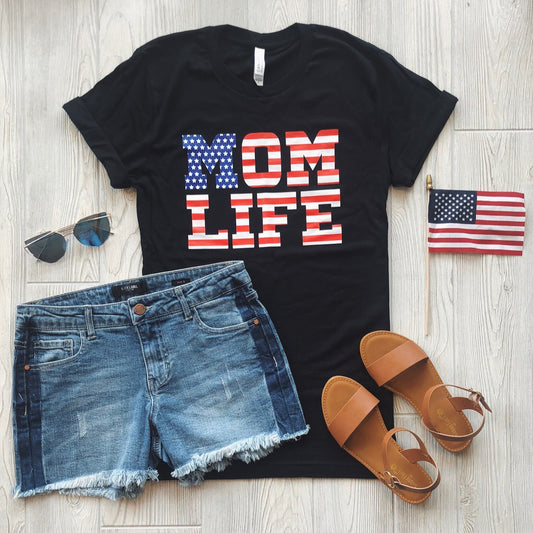 A flat lay of the Mom Flag Print • Black Tee in black, styled with denim shorts, brown sandals, sunglasses, and a small American flag on a light wooden floor.
