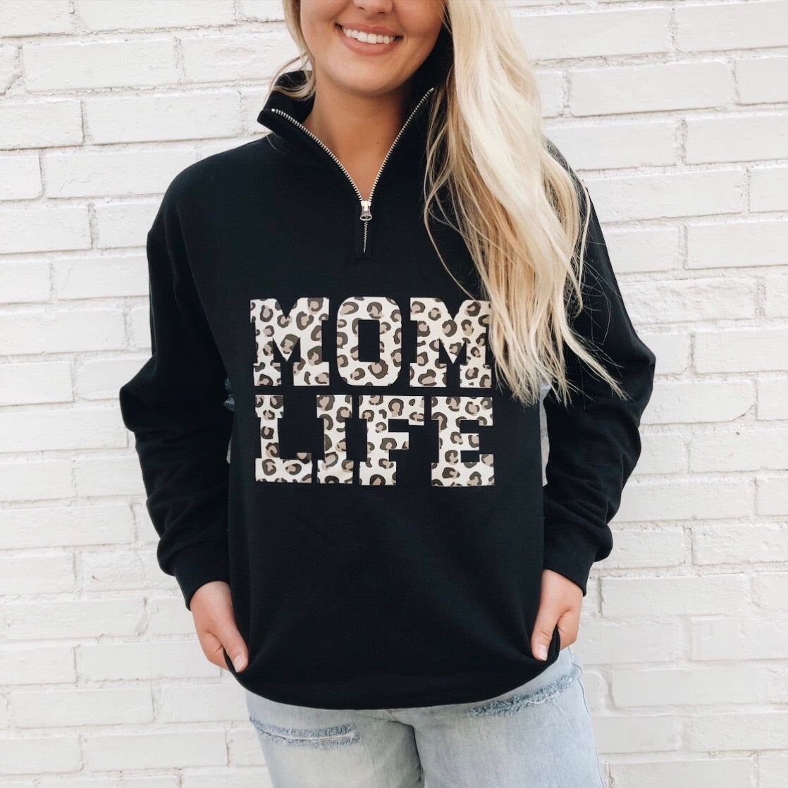 A woman with long blonde hair models the Mom Life Cheetah • Quarter Zip, featuring bold cheetah print MOM LIFE lettering on the front and a 1/4 zip design, standing against a white brick wall with her hands in her pockets.