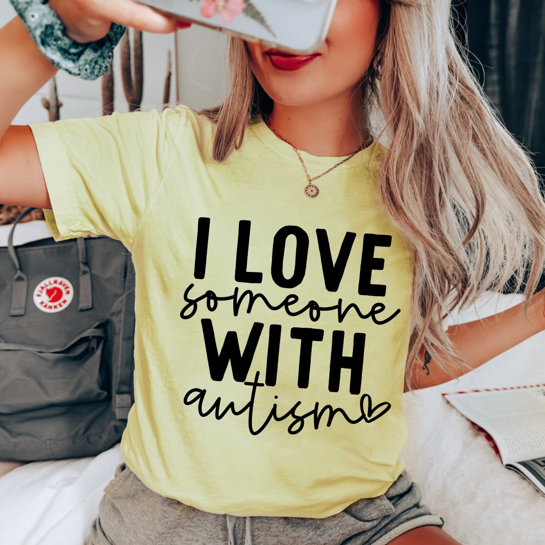 A woman with long blond hair wears the Autism Love • Yellow Tee, featuring I love someone with autism in bold black letters. Indoors, she smiles for a selfie, a gray backpack behind her. The tee has a retail fit and looks comfortable and stylish.