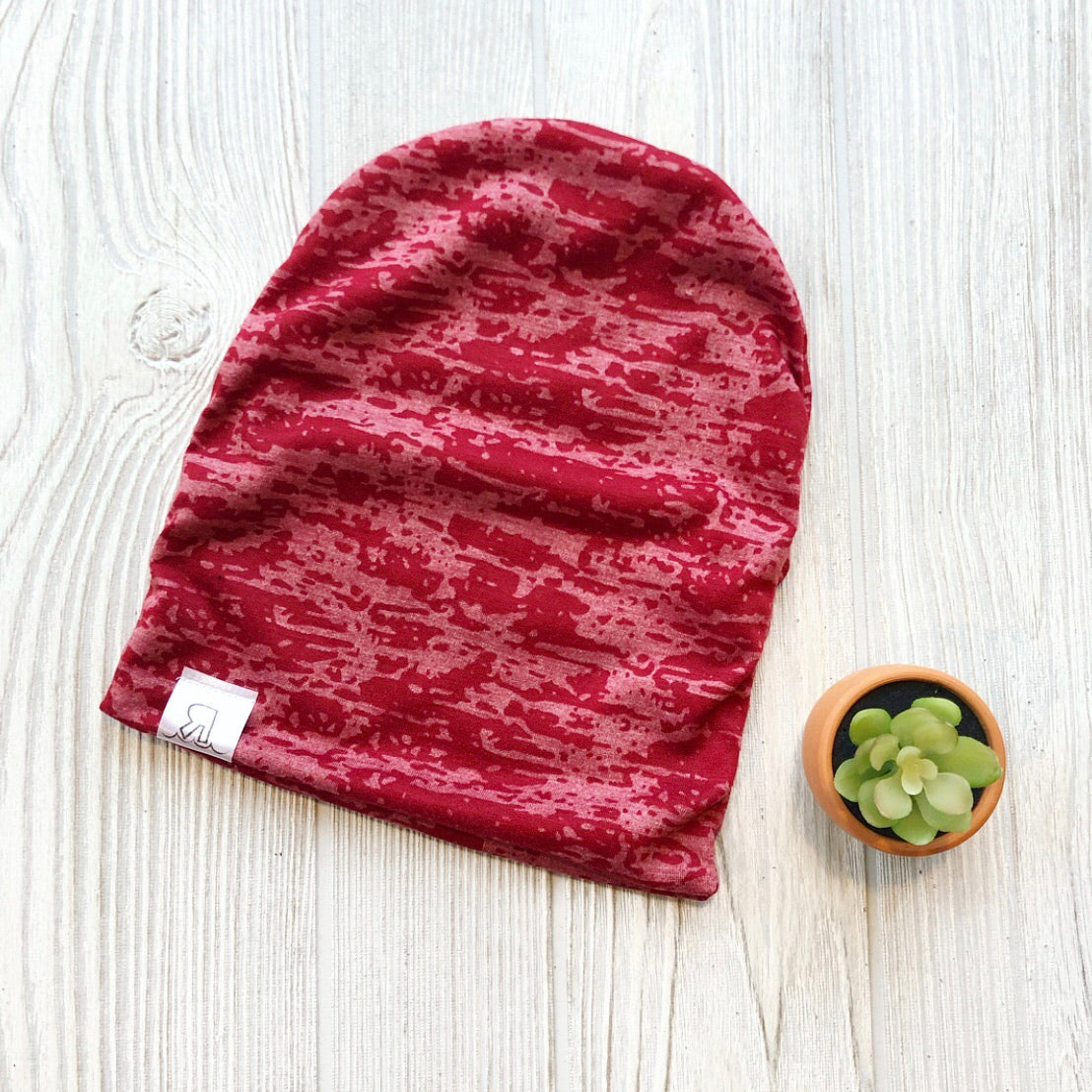 A Red Acid Wash Beanie with a small white tag is laid flat on a light wooden surface next to a small potted succulent.