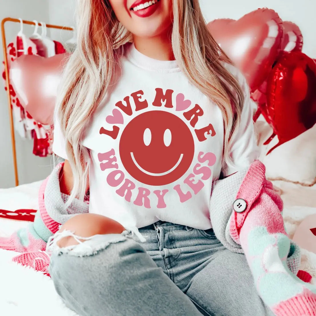 A woman sits on a bed with heart-shaped balloons, smiling in ripped jeans and a pink cardigan, wearing the Love More • White Tee featuring Love More Worry Less and a pink smiley face.