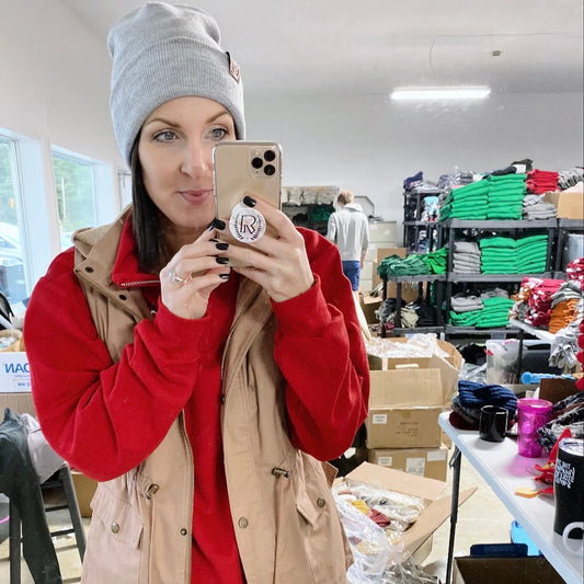 A woman wearing a gray Sunshine Collection Beanie with hand-sewn leather logo patches, a red sweater, and beige vest takes a selfie in a room filled with shelves of folded clothes and stacked boxes.