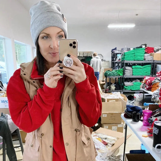 A woman wearing a gray Sunshine Collection Beanie with hand-sewn leather logo patches, a red sweater, and beige vest takes a selfie in a room filled with shelves of folded clothes and stacked boxes.