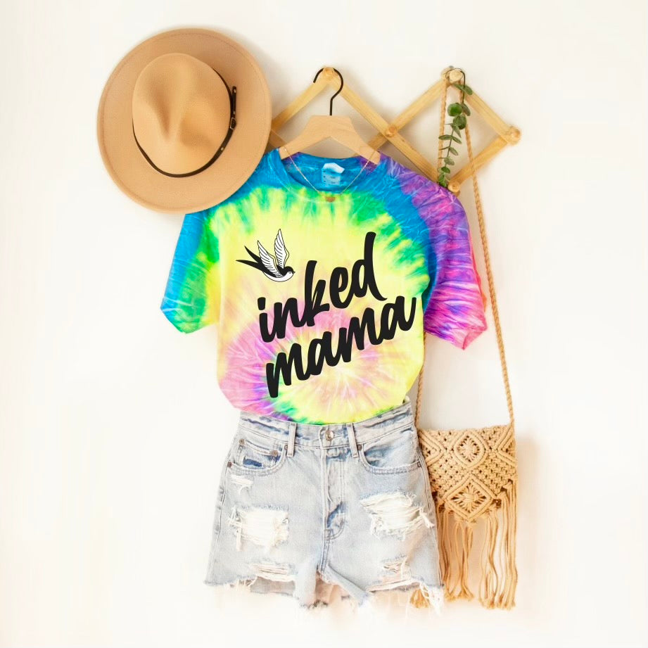 The Inked Mama • Neon Tie Dye tee features a vibrant bird graphic and hangs on a wooden rack with distressed denim shorts, a tan hat, and a woven bag—perfect for laid-back, colorful style. This tee offers a versatile unisex fit.