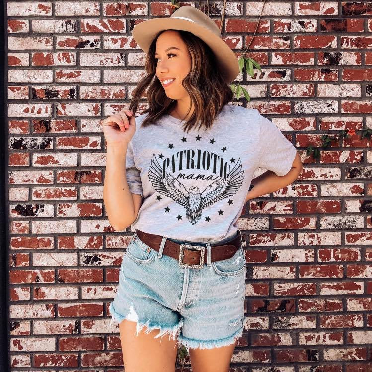 A woman in a wide-brimmed hat, Patriotic Mama Eagle Grey Tee, and denim shorts smiles in front of a brick wall, holding her hair with one hand.
