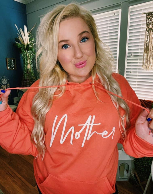 A woman with long blonde hair and blue eyes smiles indoors, holding the hoodie strings of the Mother Script • Coral Hoodie. She stands in a bright room with white blinds and plants in the background.
