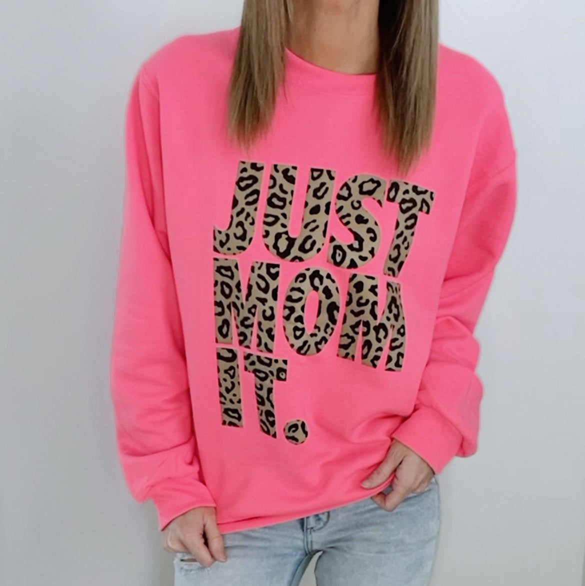 A person wears the JUST MOM IT • Neon Pink Pullover, featuring bold cheetah print lettering. The unisex fit pairs well with their straight, light brown hair and light blue jeans.