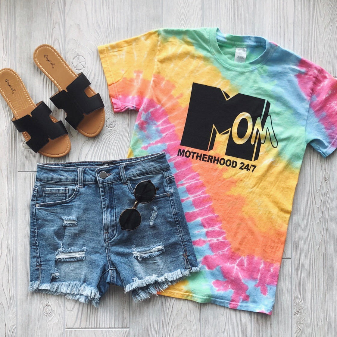 Flat lay of the Mom Life 24/7 Tie-Dye Tee, distressed denim shorts, black sunglasses, and black slide sandals on a light wood floor.