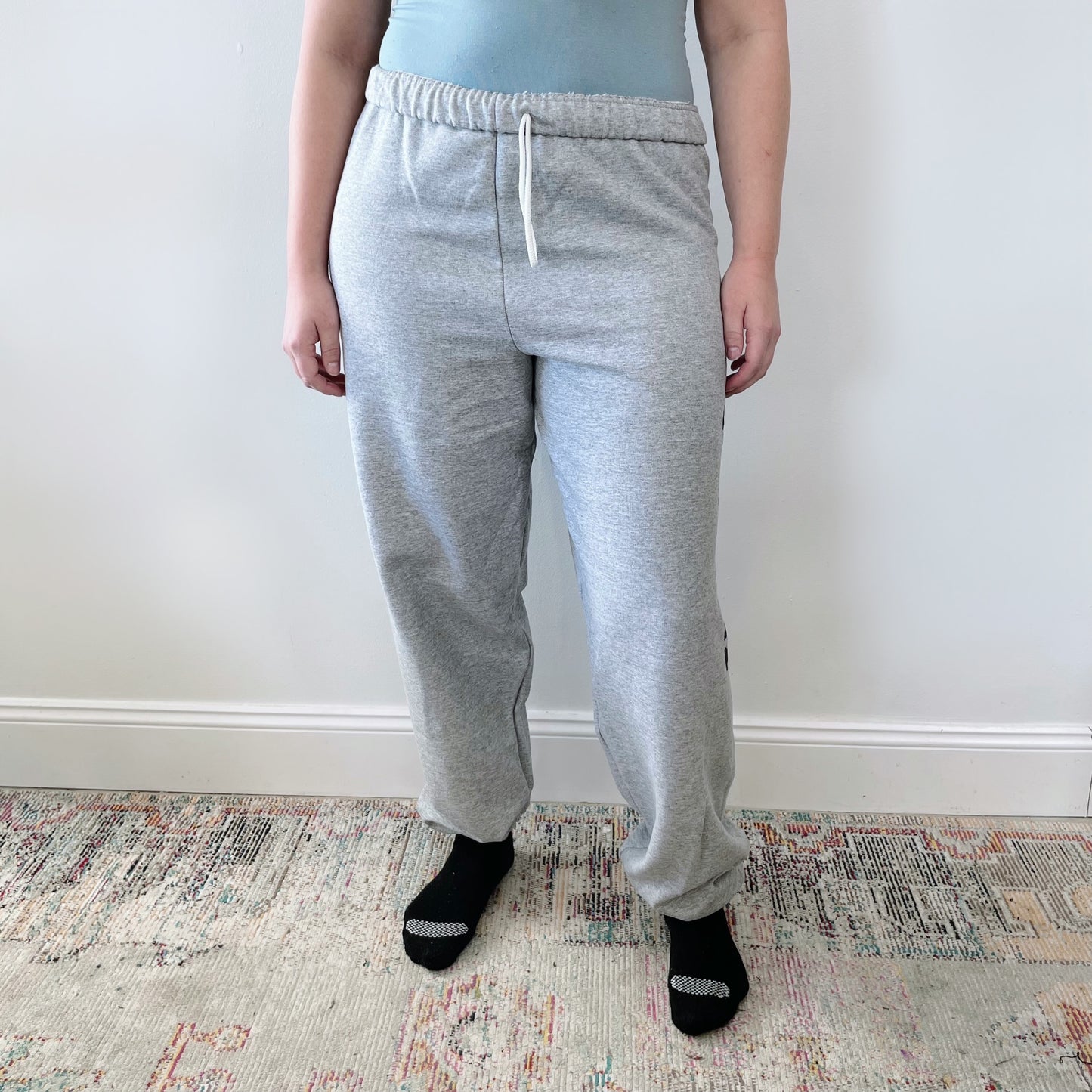 A person stands indoors against a plain light wall, wearing a light blue top, MAMA Sweats • gray cozy fleece joggers with a drawstring, and black socks. The image is cropped to show the body from shoulders to feet.