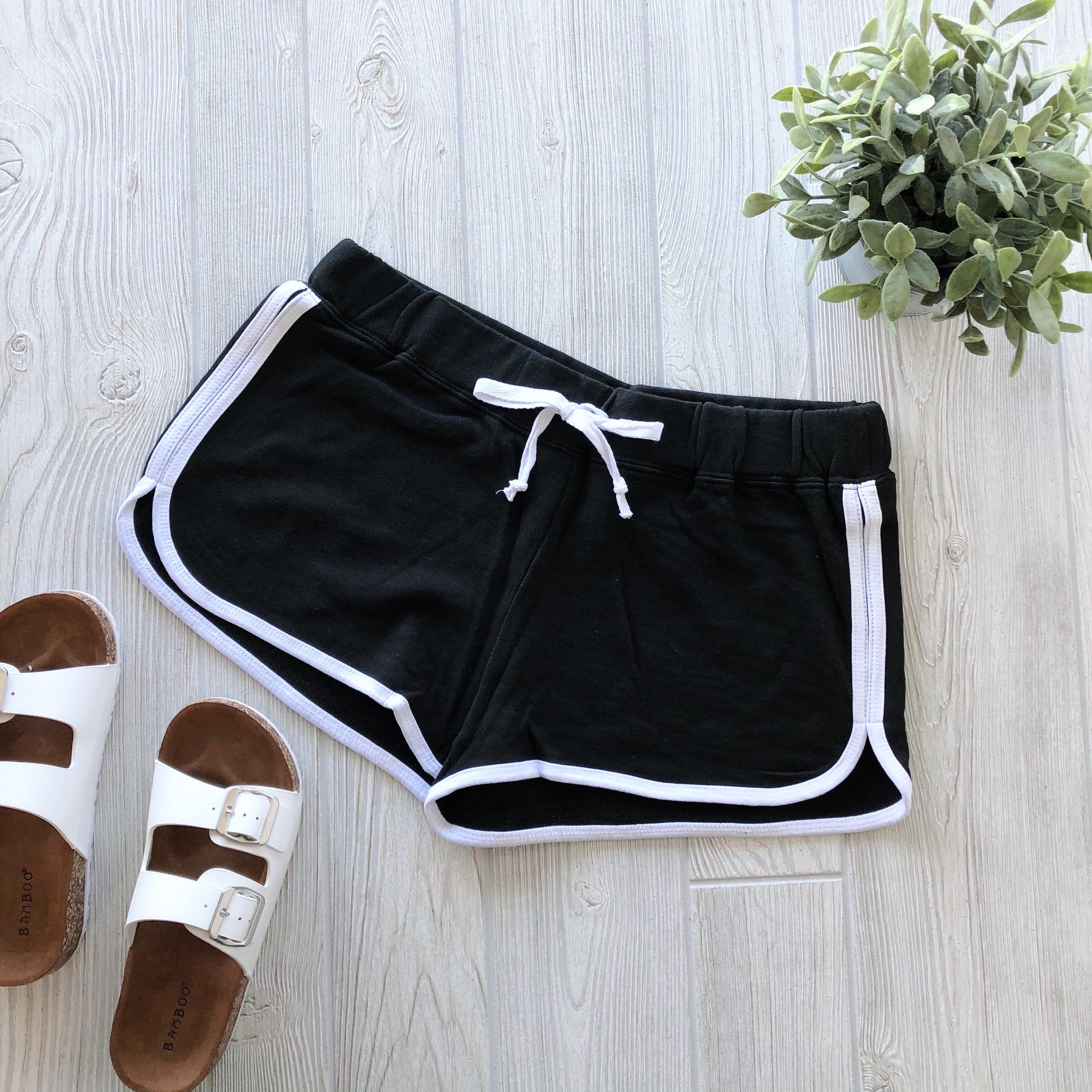 Relay Shorts • More Colors!—black vintage-style womens athletic shorts with white trim and drawstring—are displayed on a light wooden floor beside white sandals and a green potted plant.