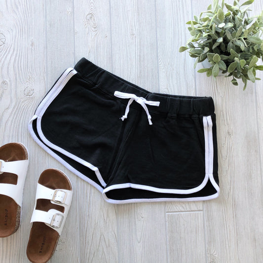 Relay Shorts • More Colors!—black vintage-style womens athletic shorts with white trim and drawstring—are displayed on a light wooden floor beside white sandals and a green potted plant.