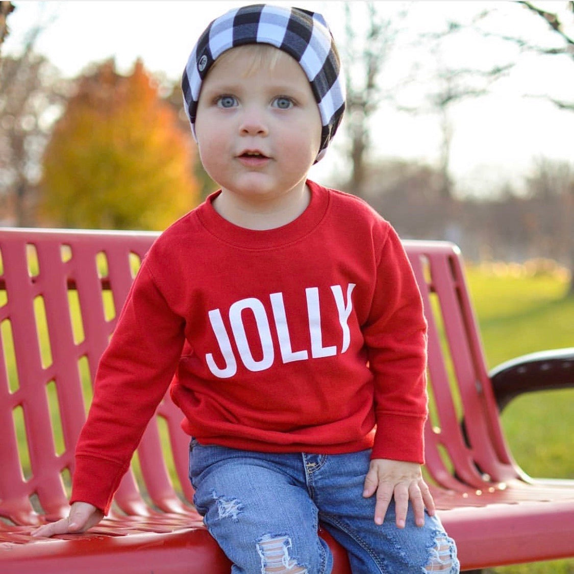 A young child wears the Jolly Pullover • Kids, ripped jeans, and a black-and-white checkered beanie while sitting on a red bench outdoors, surrounded by green grass and trees.