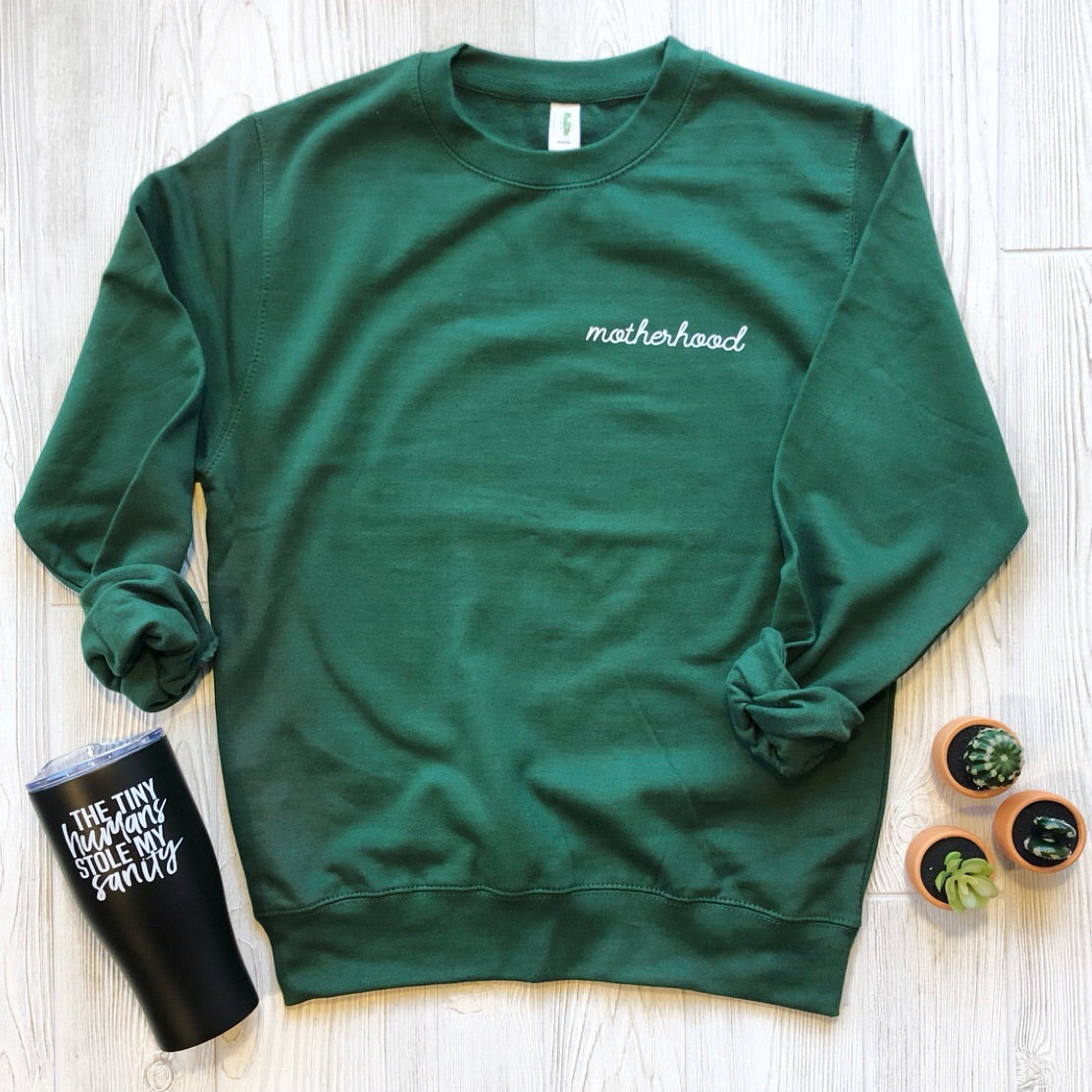 The Motherhood Left Chest • Forest Pullover, a forest green fleece sweatshirt with white embroidery, is displayed on a white wood surface beside a black tumbler with white text and two small potted succulents.