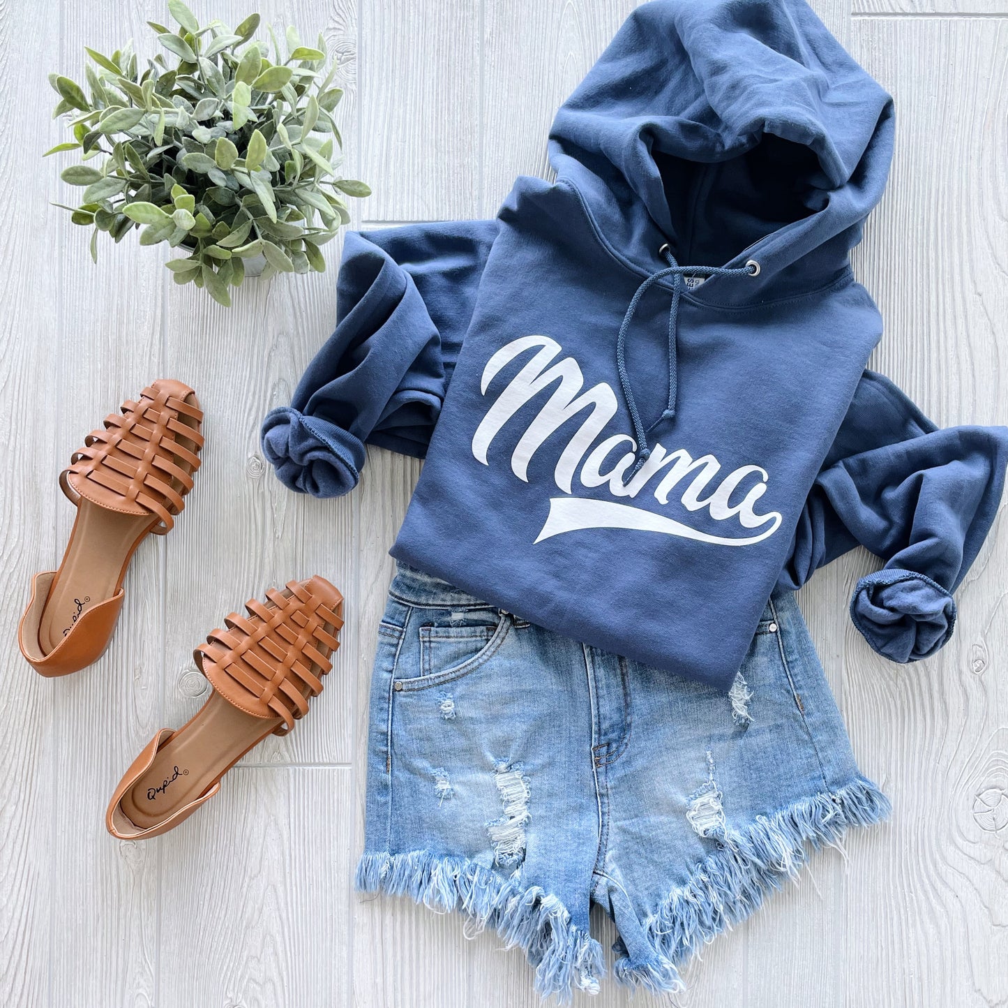 The Mama Sporty • Denim Hoodie—a blue unisex fleece sweatshirt with Mama in white script—is styled with distressed denim shorts, brown woven sandals, and a green potted plant on a light wooden floor for cozy bonfire vibes.