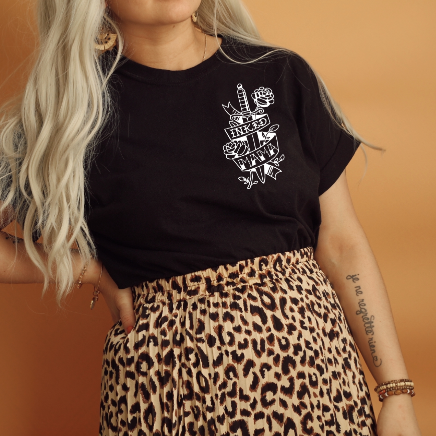 A woman with long, wavy platinum blonde hair models the Inked Mama • Black Tee, featuring a white tattoo-style graphic, paired with a leopard print skirt and accessories, standing against a tan background.