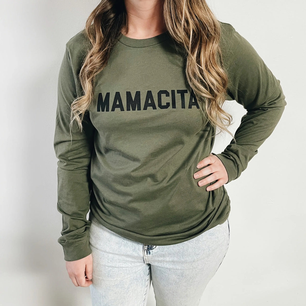 A woman with long wavy hair models the Mamacita • Olive Long Sleeved tee in unisex sizing, featuring bold black MAMACITA lettering. She pairs it with light blue jeans and poses with one hand on her hip against a plain background.