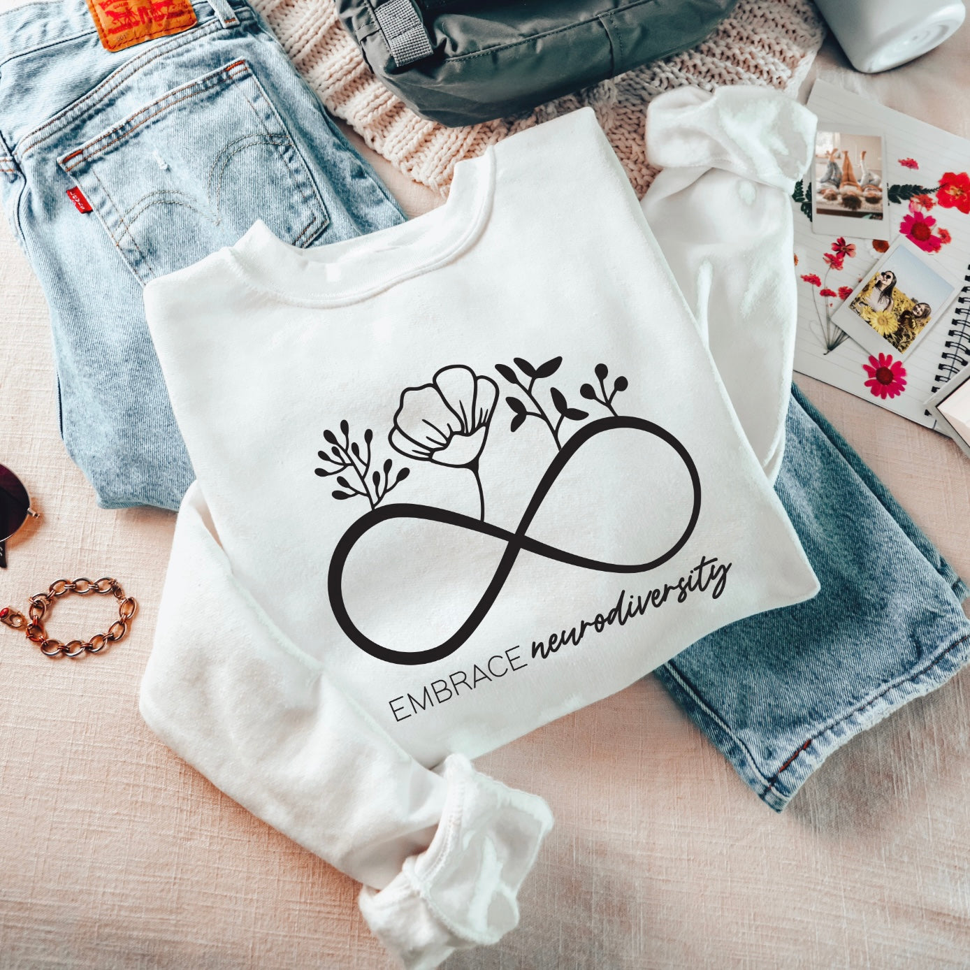 The Embrace Neurodiversity • White Pullover, a cozy unisex sweatshirt with an infinity symbol and floral accents, is folded on a bed beside blue jeans, a gray backpack, a gold bracelet, and floral-themed accessories.