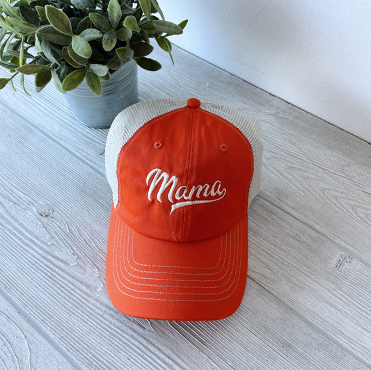 The Sporty Mama Mesh Hat in orange, a unisex cap with Mama embroidered on the front, rests on a light wooden surface next to a small potted plant with green leaves.