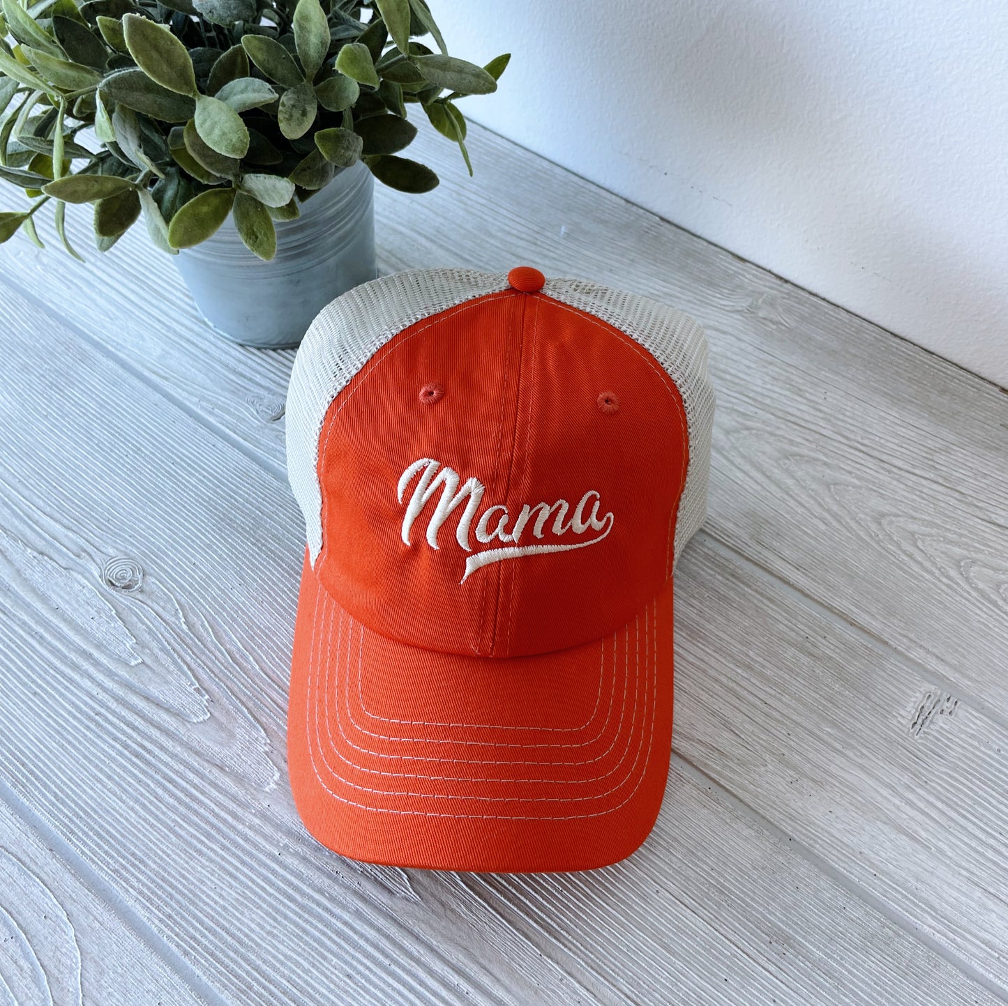 The Sporty Mama Mesh Hat in orange, a unisex cap with Mama embroidered on the front, rests on a light wooden surface next to a small potted plant with green leaves.