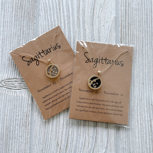 Two Zodiac Sign • Necklace pieces for Sagittarius with adjustable clasps are displayed on gold chains over brown zodiac description cards, each sealed in clear plastic and set on a light wood surface.