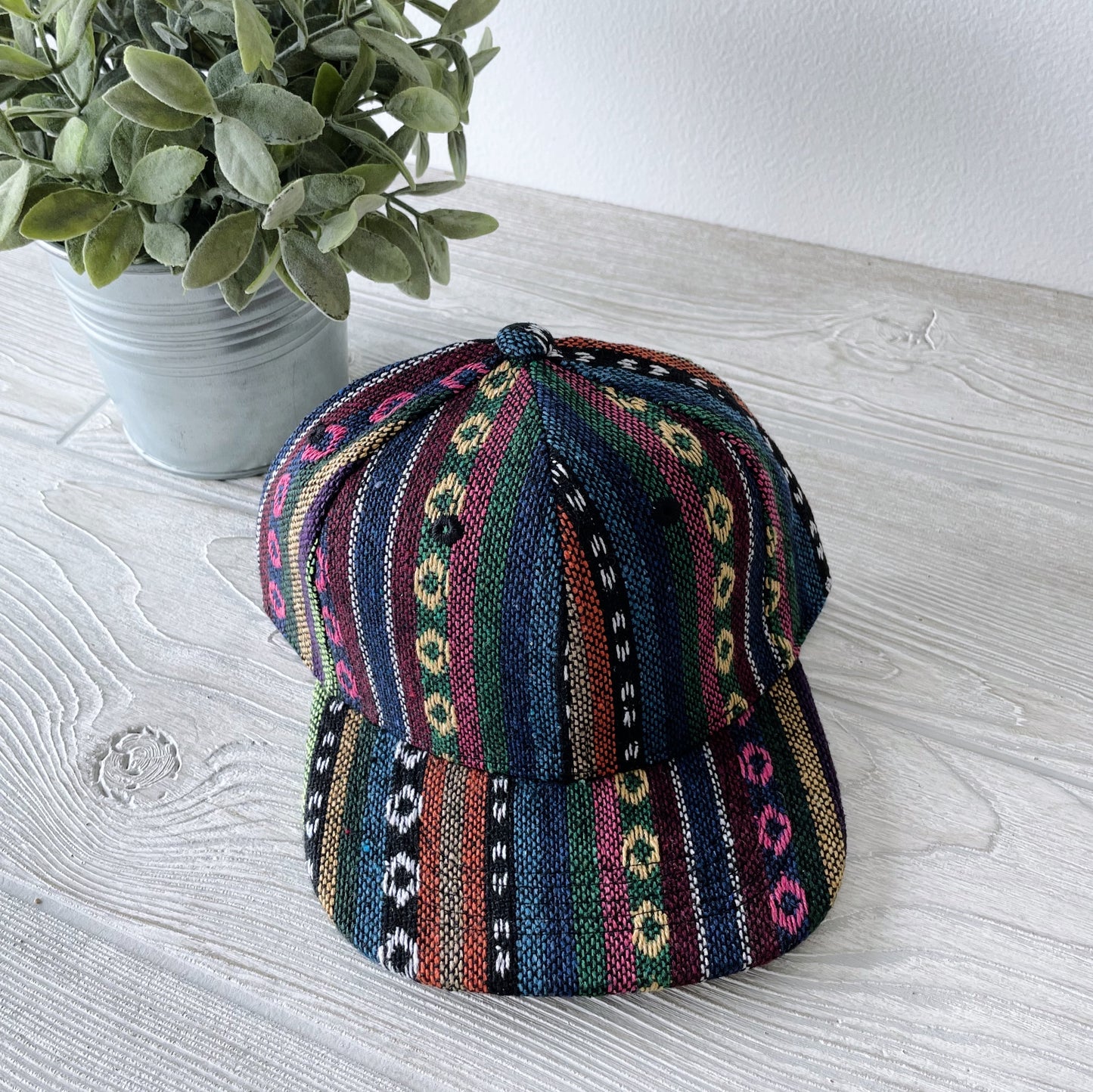 A Kids Snapback • Orlo featuring vibrant vertical stripes and geometric patterns rests on a light wood surface beside a potted green plant.