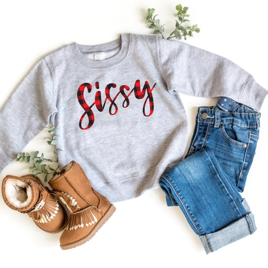 The Sissy Plaid Pullover, featuring Sissy in bold red buffalo plaid, is styled with blue jeans and tan moccasin boots on a white background, accented by green sprigs.