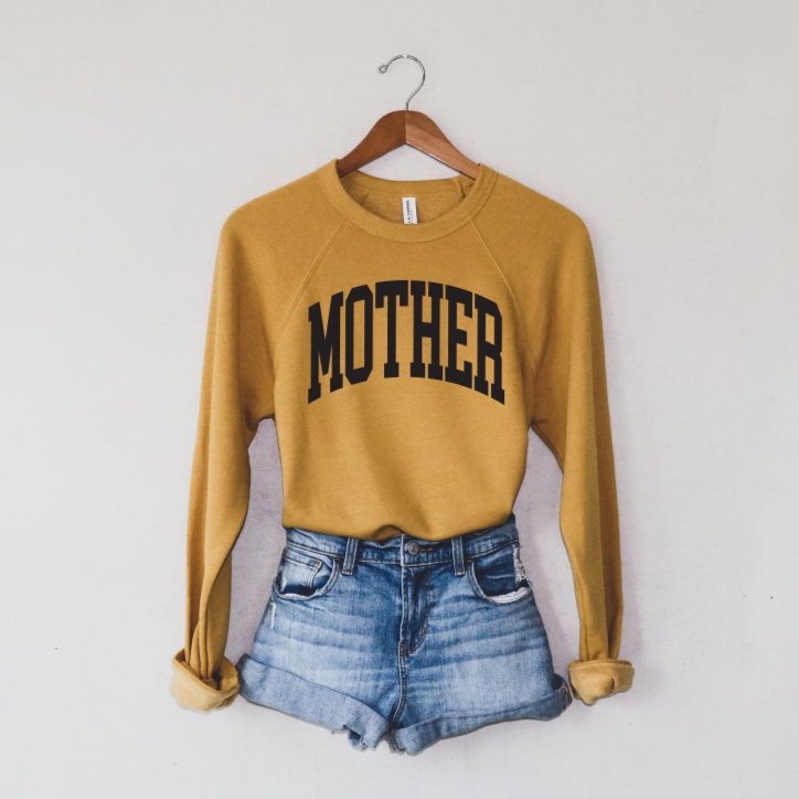 The MOTHER • Mustard Pullover, featuring a bold college design, hangs on a wooden hanger next to rolled-up denim shorts against a plain white wall.