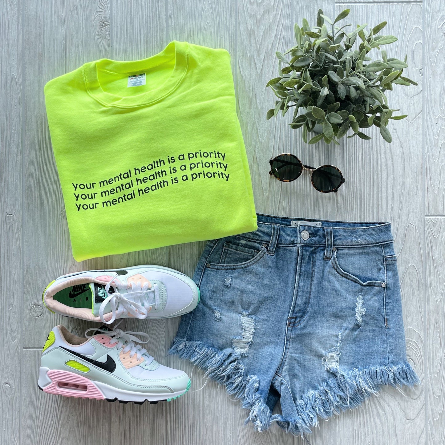 Neon yellow Mental Health • Neon Yellow Pullover sweatshirt, unisex fit, paired with distressed denim shorts, pastel-accented white Nike sneakers, round sunglasses, and a small potted plant on a light wood floor.