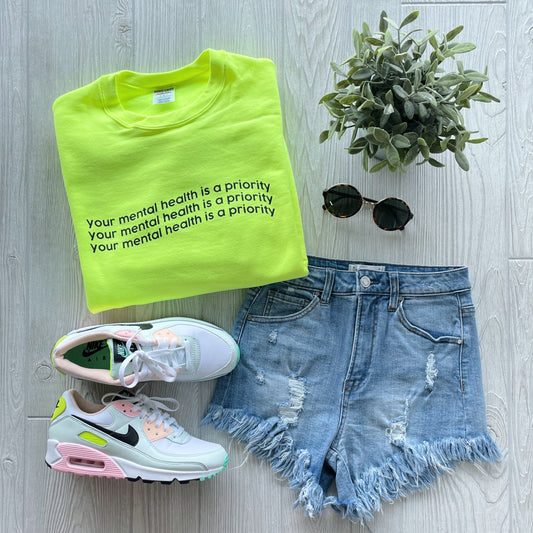 Neon yellow Mental Health • Neon Yellow Pullover sweatshirt, unisex fit, paired with distressed denim shorts, pastel-accented white Nike sneakers, round sunglasses, and a small potted plant on a light wood floor.