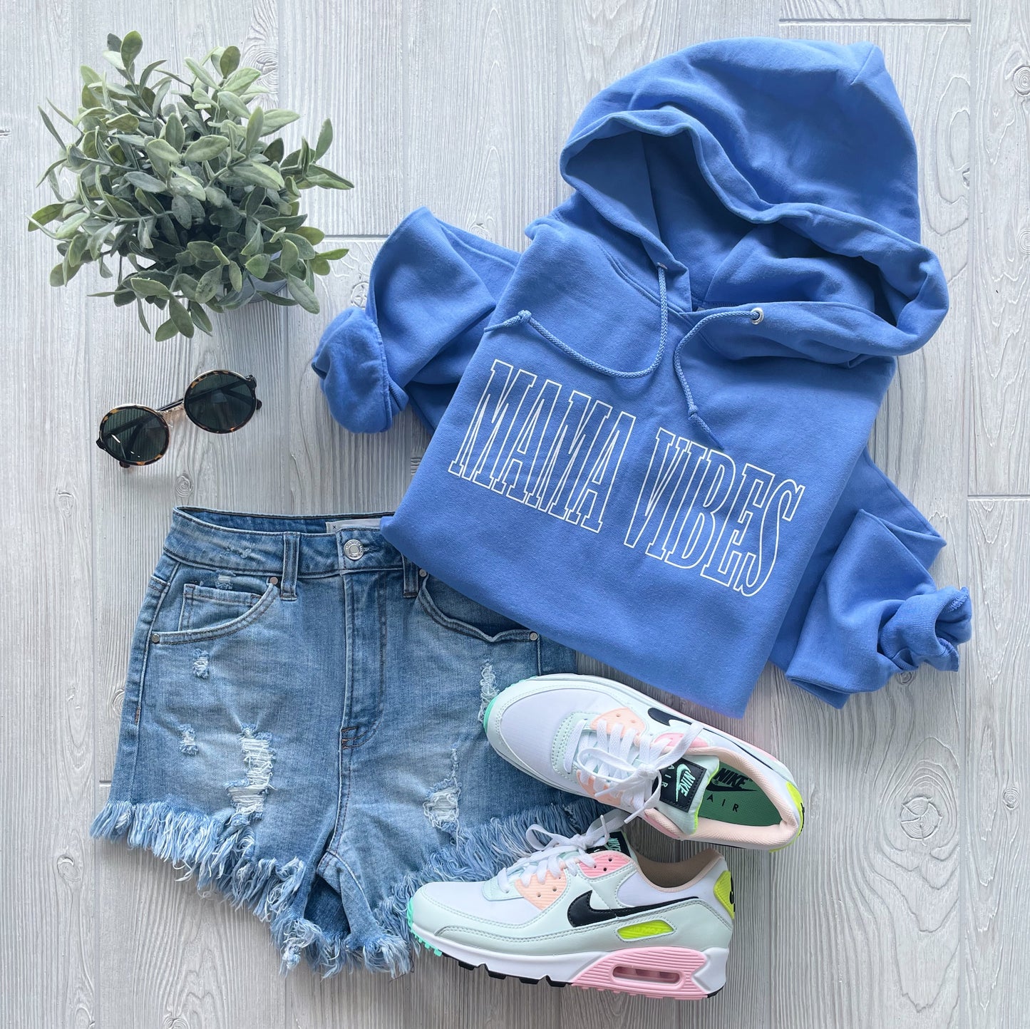 A flat lay of the MAMA VIBES • Periwinkle Hoodie made with pill-resistant fleece, distressed denim shorts, white Nike sneakers with colorful accents, black sunglasses, and a green potted plant on a light wood surface.