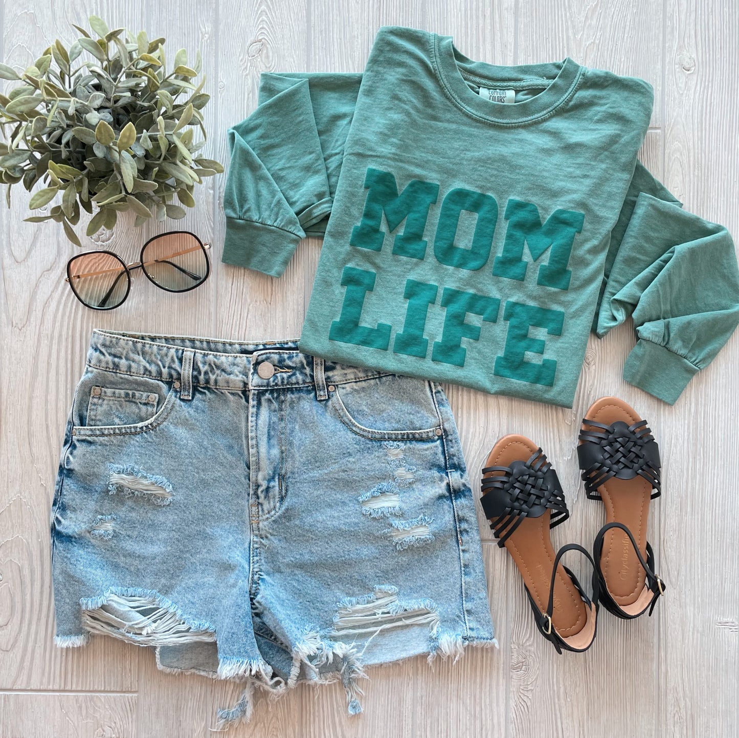 Flat lay of the MOM LIFE teal puff • Comfort Long Sleeve tee, crafted from 100% ring-spun cotton, paired with distressed denim shorts, black sandals, round sunglasses, and a small potted plant on a light wood background.