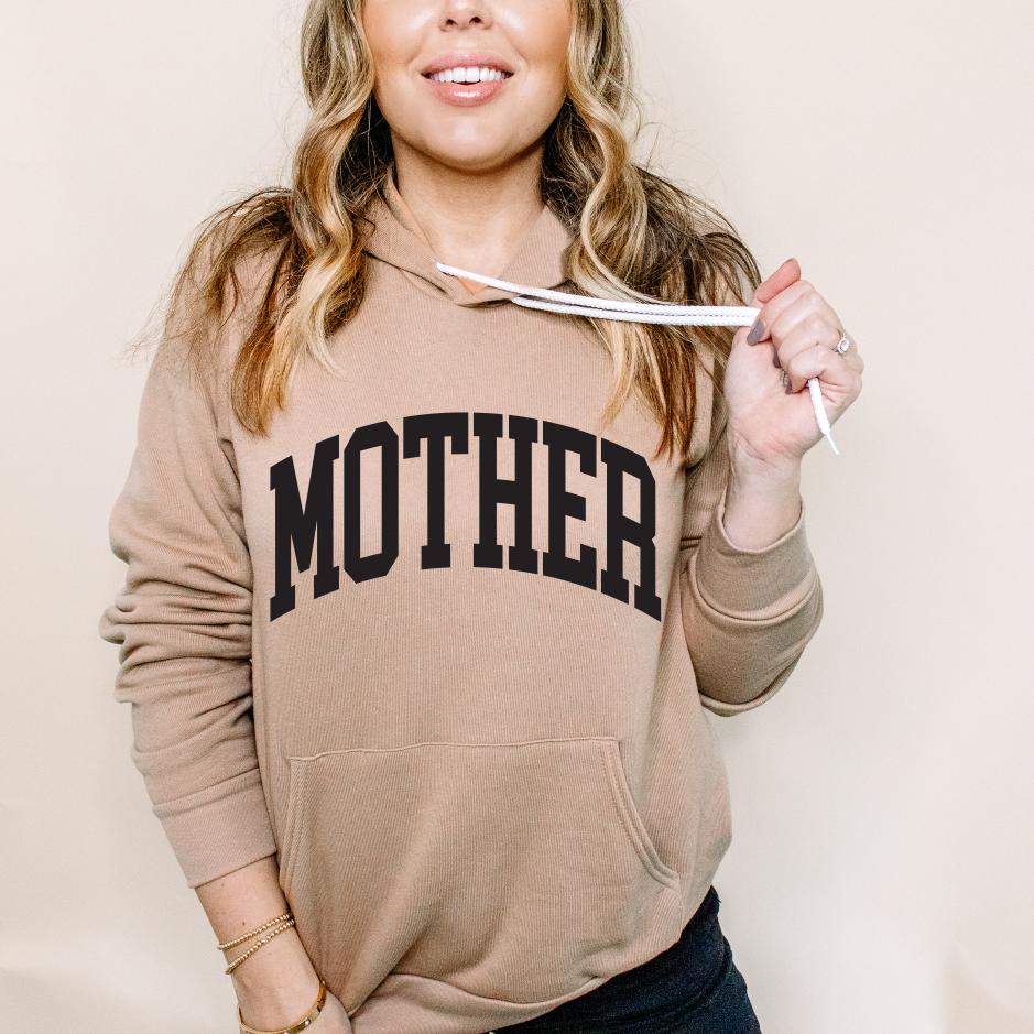 A woman smiles against a plain light background, holding the strings of the MOTHER • Sand Hoodie—a unisex beige hoodie with a bold MOTHER design on the front, made from soft sponge fleece fabric.