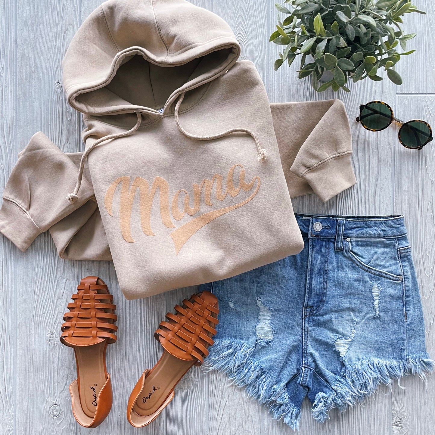 Flat lay of the Tonal Sporty Mama • Hoodie in beige, a unisex heavyweight style made with cozy 3-end fleece, paired with distressed denim shorts, brown woven sandals, round sunglasses, and a small potted plant on a light wooden surface.