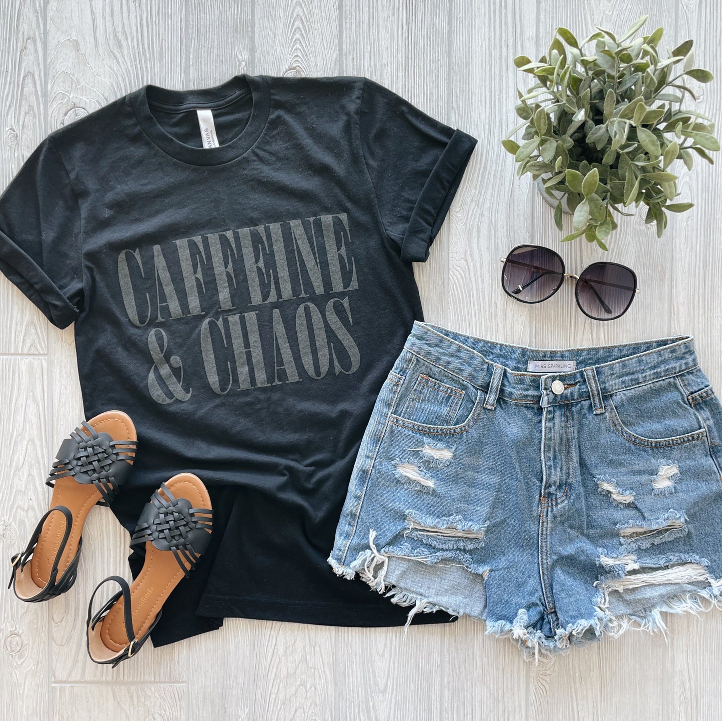 Flat lay of a black Caffeine and Chaos • Tonal Tee with a unisex fit, paired with distressed denim shorts, black sandals, sunglasses, and a potted green plant on a light wooden surface.