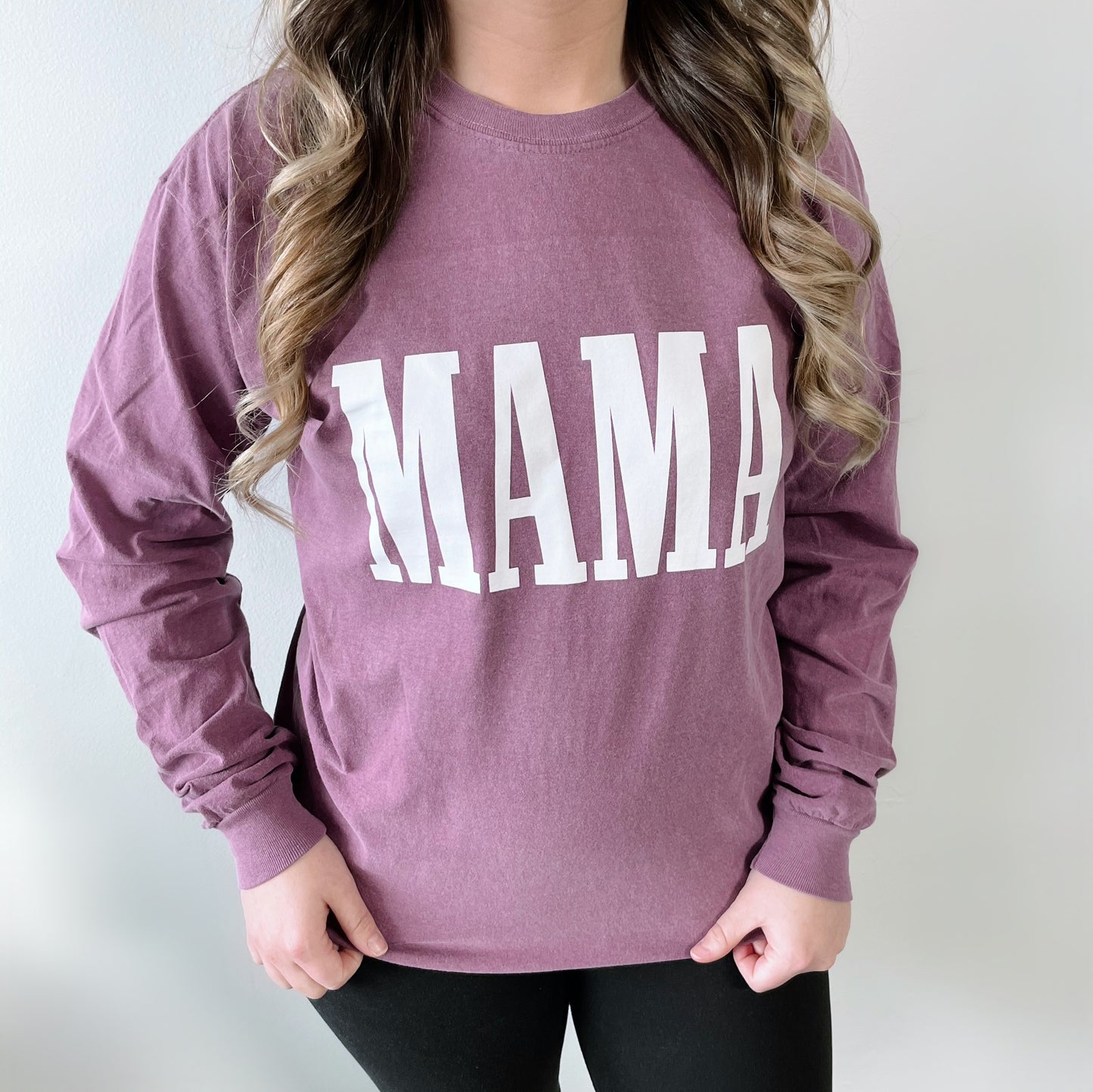 A person wears the MAMA • Plum Long Sleeve, featuring MAMA in bold white letters on the front, standing against a plain light background.