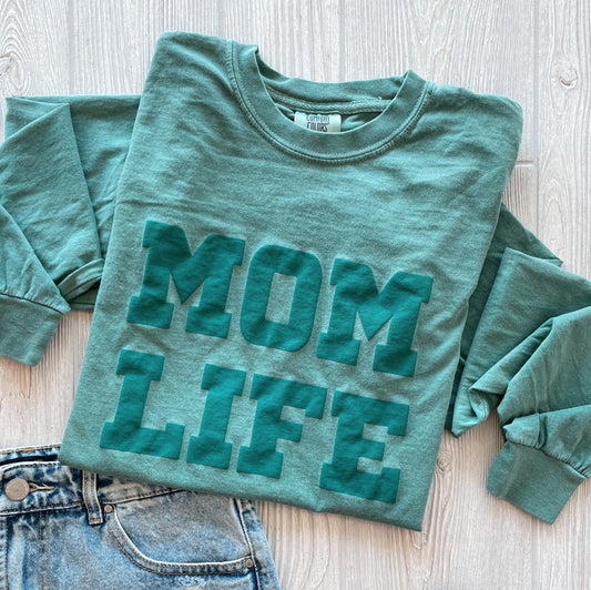 The MOM LIFE teal puff • Comfort Long Sleeve, crafted from 100% ring-spun cotton with bold raised MOM LIFE lettering, is folded on a light wood surface beside blue denim jeans.