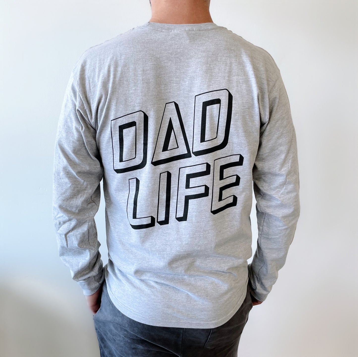 A person wears the Skater Dad • Long Sleeve, a light gray crew neck shirt made in Michigan with bold black DAD LIFE text on the back, where the A in DAD is stylized as a triangle.