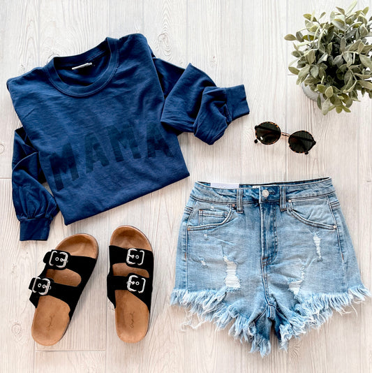 A flat lay of a casual outfit with the MAMA • Tonal Navy Long Sleeve, distressed denim shorts, black sandals, round sunglasses, and a small potted plant on a light wood surface.