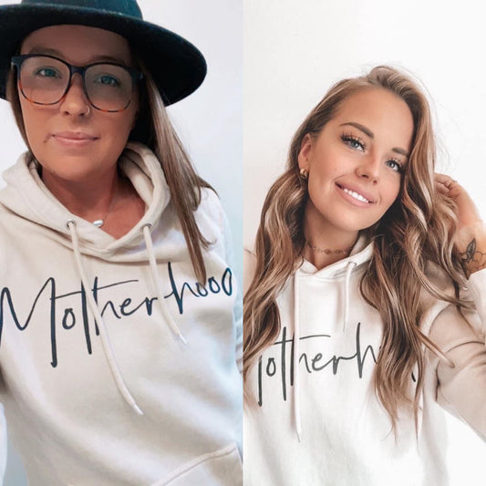 Two women wear the Motherhood • Sand Hoodie with Motherhood in cursive; one on the left has glasses and a black hat, while the other smiles, touching her hair. This cozy hoodie offers a unisex fit.