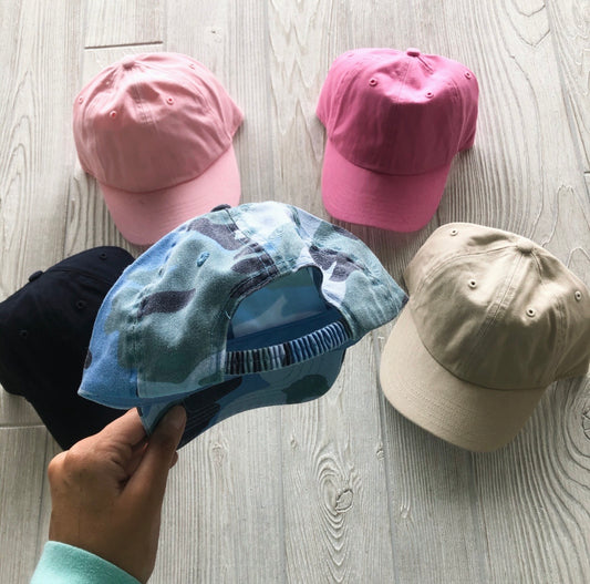 A hand holds a blue camo Kids Baseball Hat • More Colors! above four other caps—black, pink, tan, and pink—all with elastic bands for a perfect toddler fit, arranged on a light wooden floor.