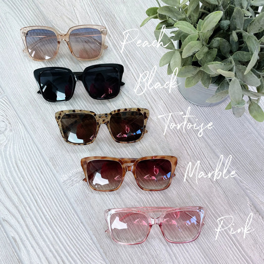 Five pairs of Natalie • Adult Sunglasses with UV400 Protection—peach, black, tortoise, marble, and pink—are vertically arranged on a light wood surface next to a green potted plant. Each pair is script-labeled. Final Sale.