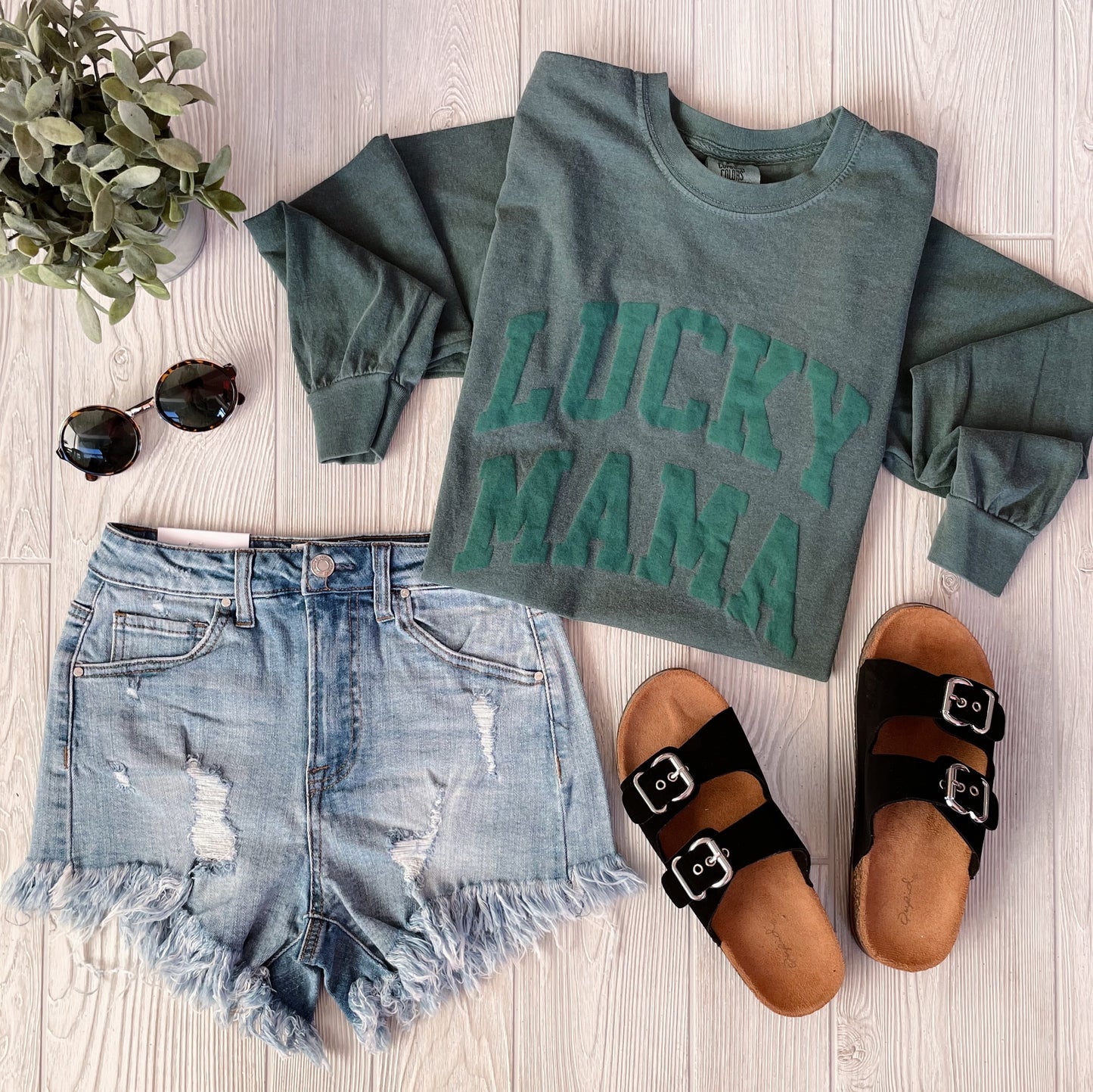 Flat lay of the LUCKY MAMA • Tonal Green Long Sleeve with garment-dyed fabric, distressed denim shorts, black buckle sandals, round sunglasses, and a small potted plant on a light wood surface.
