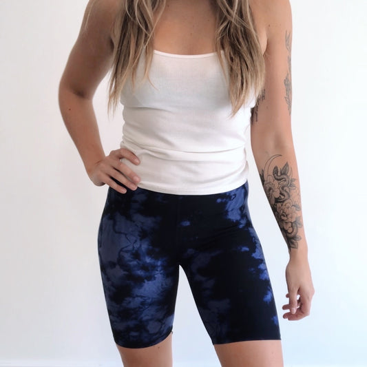 A person wearing Blue Dye • Brushed Biker Shorts, made from super-soft brushed jersey fabric, stands against a plain white background with one hand on their hip and a floral tattoo visible on their left arm.