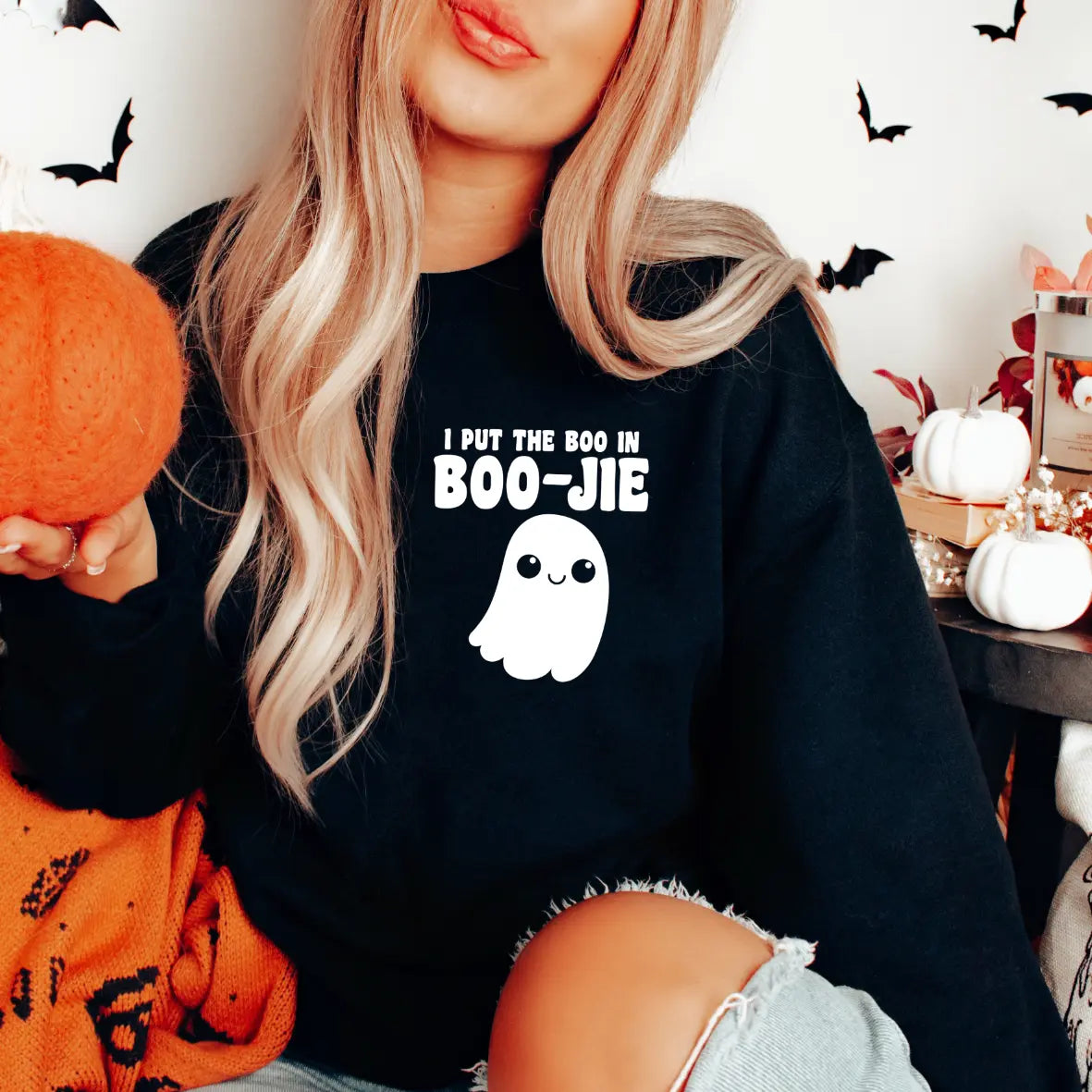 A woman with long blonde hair wears the Boo in BOO-JIE black sweatshirt, featuring a cute ghost design. She holds an orange pumpkin and is surrounded by Halloween décor like bat cutouts and small pumpkins.