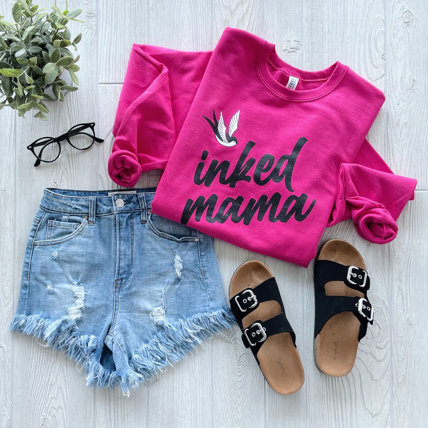 Flat lay featuring the Inked Mama • Pink Sweatshirt, distressed denim shorts, black buckle slide sandals, black eyeglasses, and a potted plant on a light wood background.